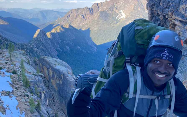 a man in mountaineering gear smiles with mountainous scenery in the background 